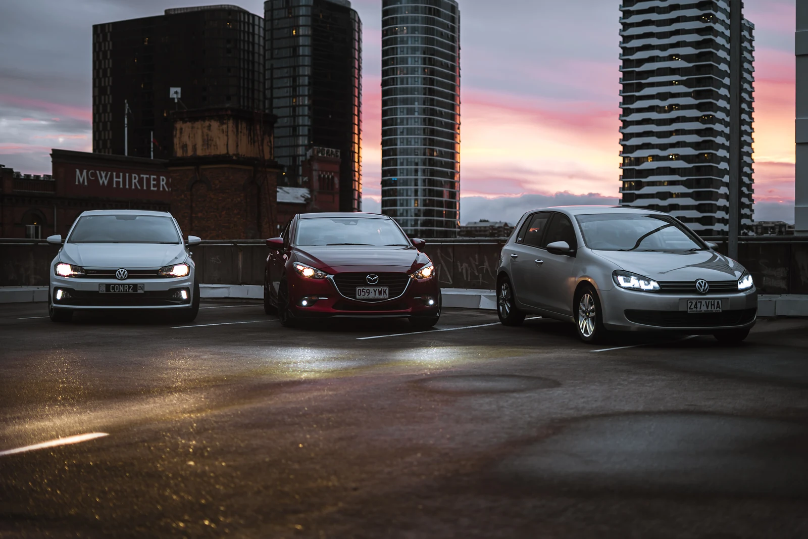 Three hatchbacks on a rooftop car park under a pink sunset sky with towers behind
