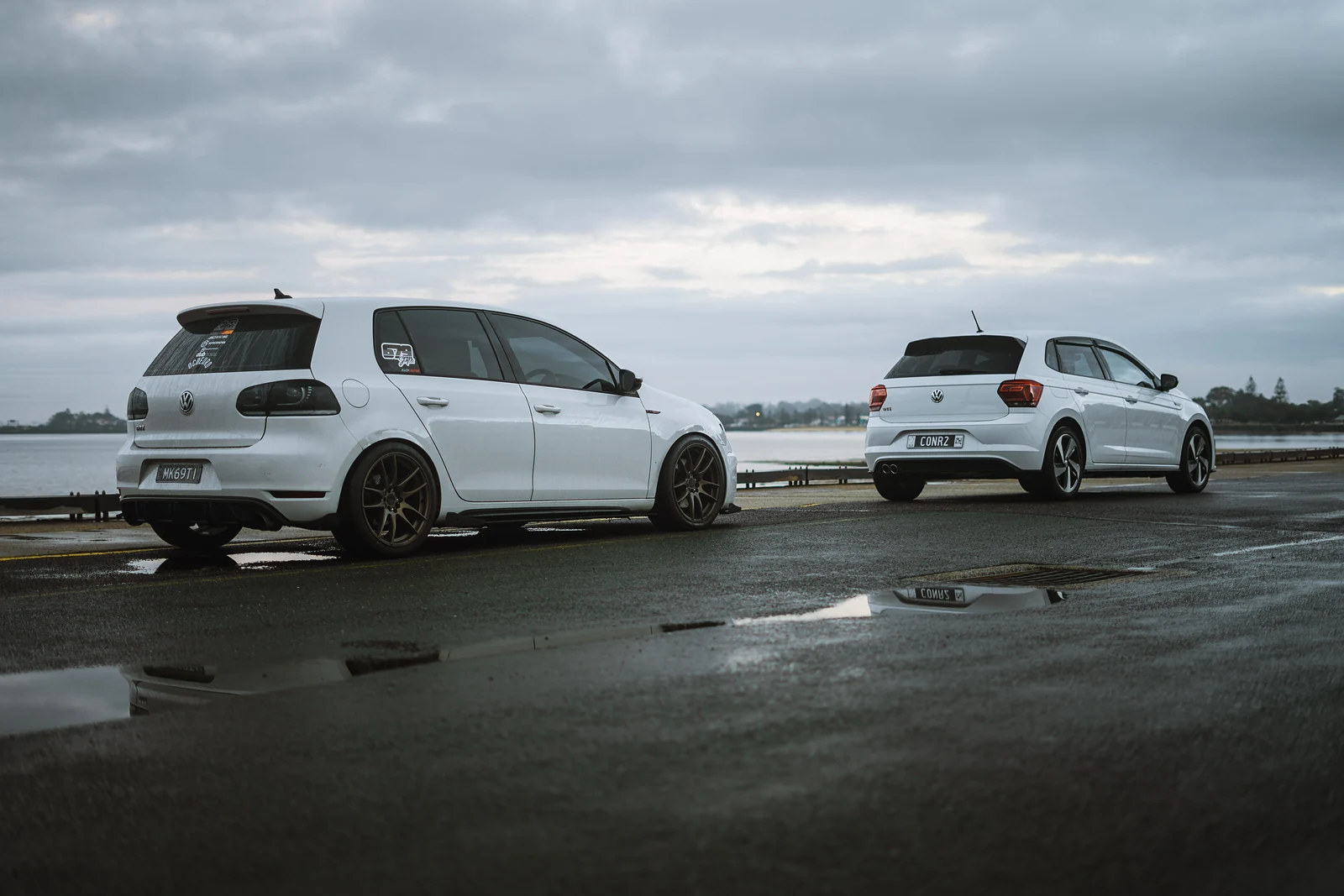Two white Volkswagen Golfs parked by the water after rain, reflected in the wet bitumen