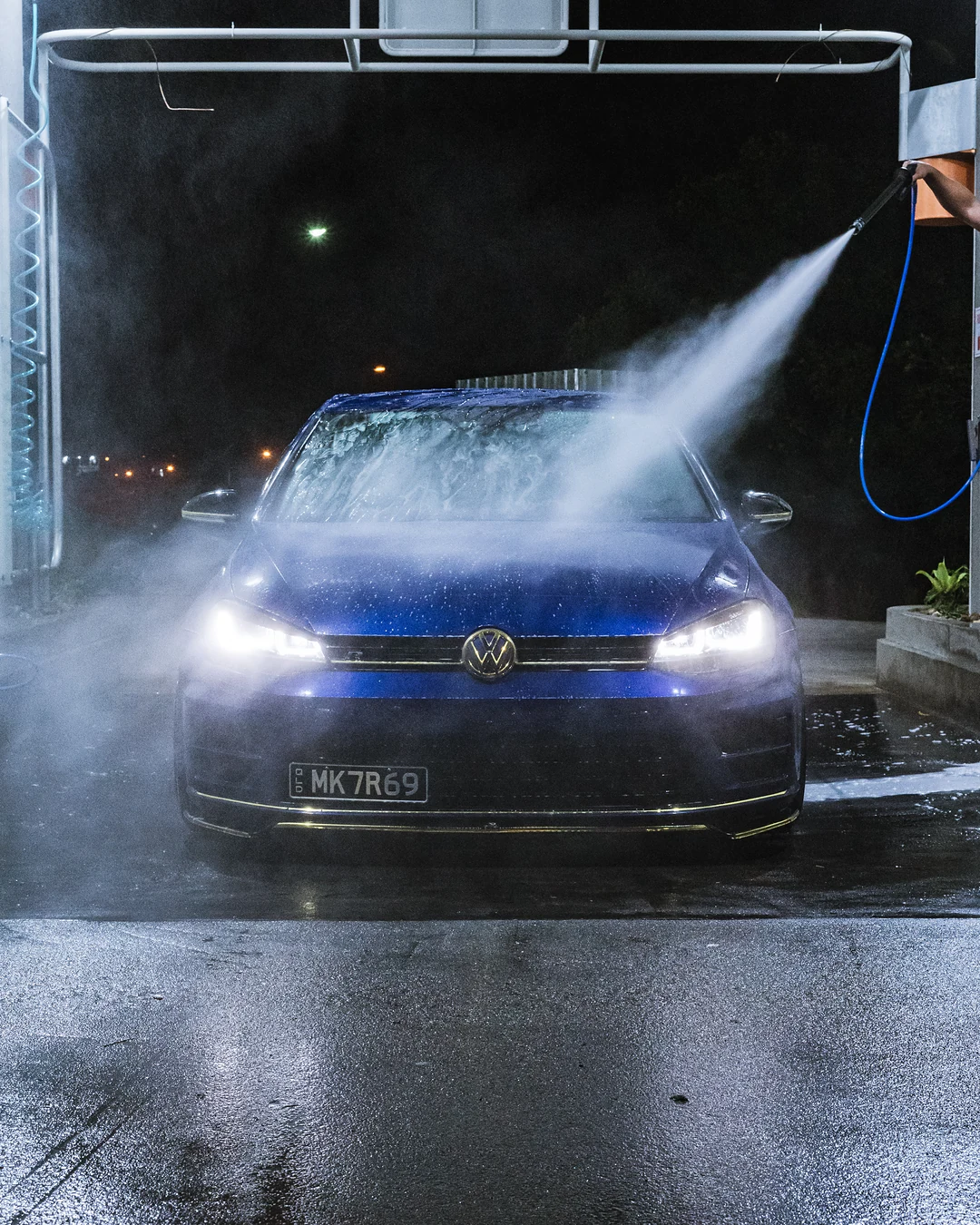 Blue Volkswagen Golf being sprayed in a wash bay at night, headlights on