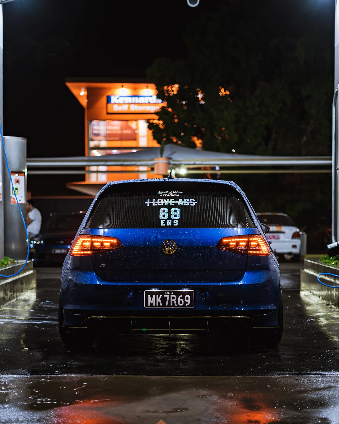 Blue Volkswagen Golf from behind in a wash bay at night, wet paint and orange lights