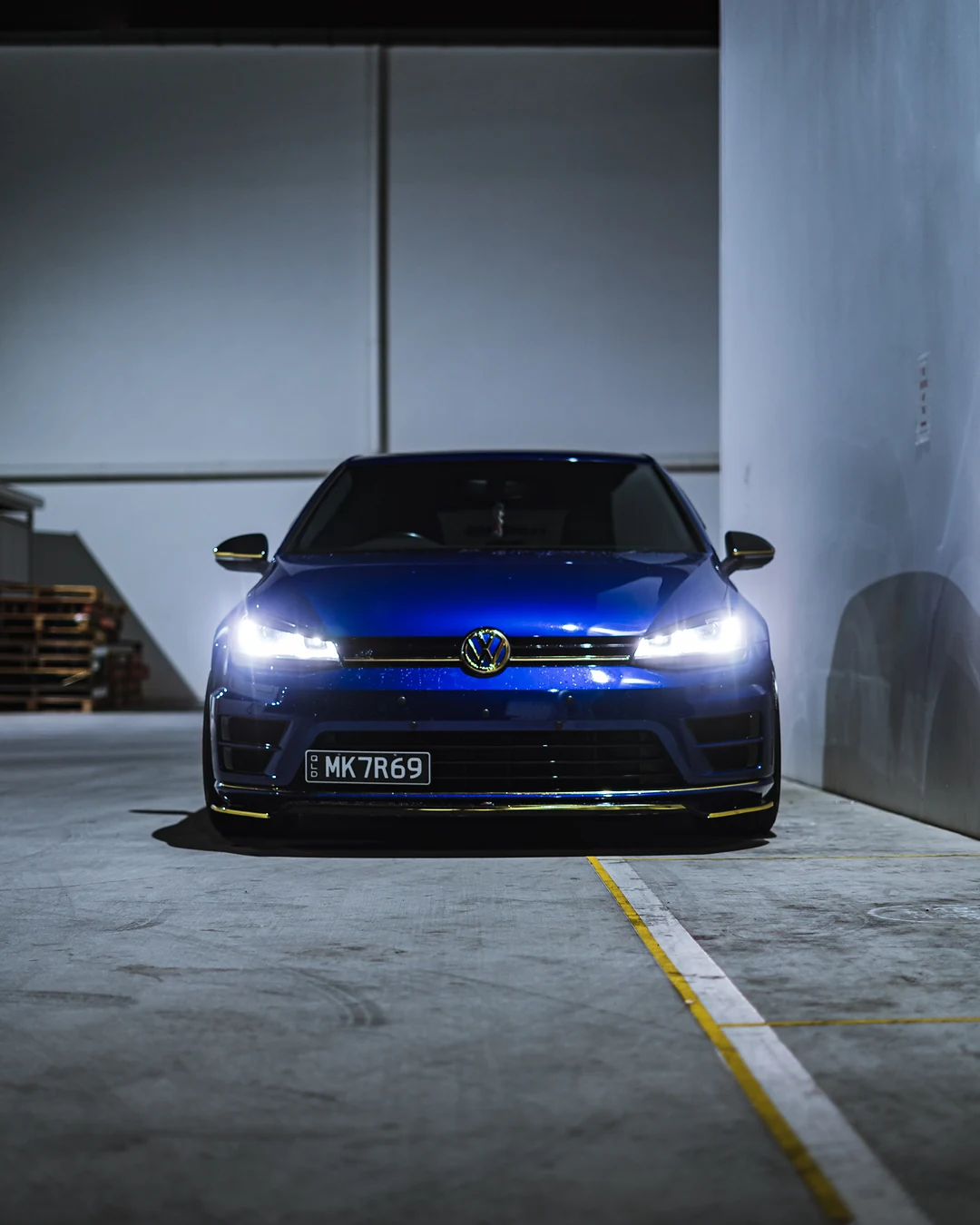 Blue Golf R with its headlights on in a white concrete car park