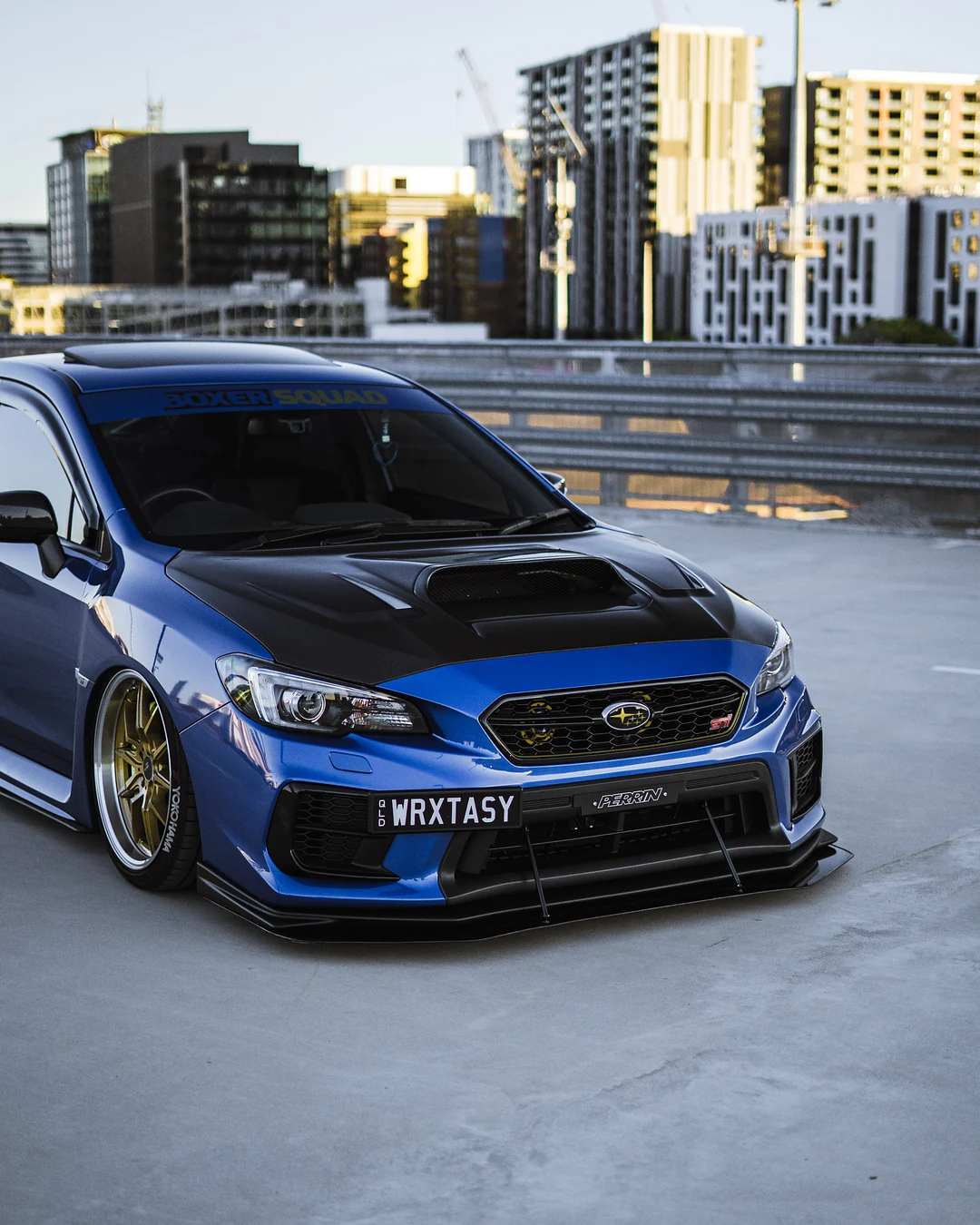 Blue Subaru WRX STI front three-quarter on a rooftop car park with city buildings behind